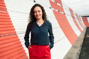 Woman with curly hair in polka dot blouse and red pants stands smiling by a red-and-white striped wall outdoors, embodying vibrant confidence akin to a leader in technology governance.