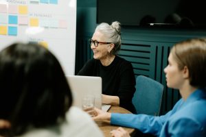 Smiling woman with gray hair sits at a desk with coworkers, laptop, and sticky notes on a board behind her, reflecting a sense of community and belonging in the workplace.