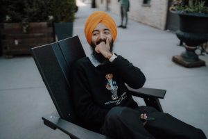Man in orange turban sitting outdoors, smiling with hand over mouth, holding a phone in one hand, radiating a sense of belonging.