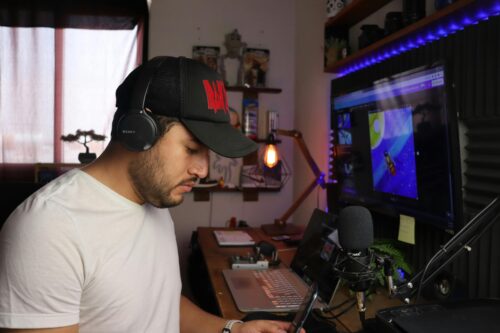In a home studio, a man wearing headphones works at his computer with a microphone, possibly engaged in crafting content for the Reporters in Residence program.