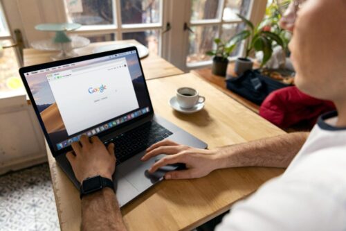 A person sits at a wooden table with a sleek MacBook, Google search open, and a steaming coffee cup beside them. Nearby plants add a touch of nature to their workspace. In the background