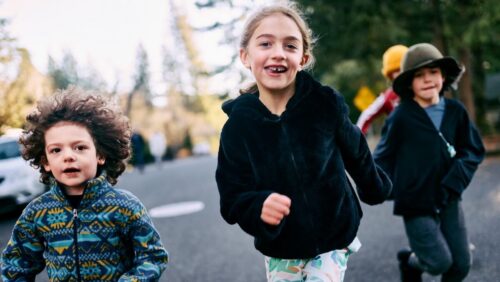 Three children, smiling as they run along the road, are framed by lush trees and a distant friend, embodying the joy of play under natures watch.