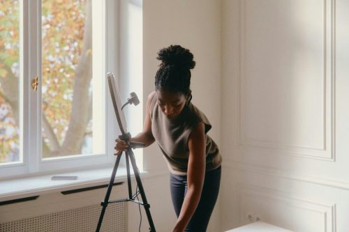 Woman adjusting a tablet on a tripod near a window in a bright room with white walls, embracing workplace innovation and modern tech culture.