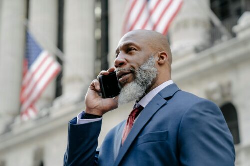 A business professional in a suit talks on his phone outside, surrounded by American flags fluttering in the breeze.