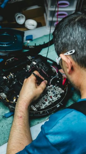A person wearing glasses repairs an electronic circuit board with tools on a workbench, exemplifying hands-on involvement in the Governance of Tech.