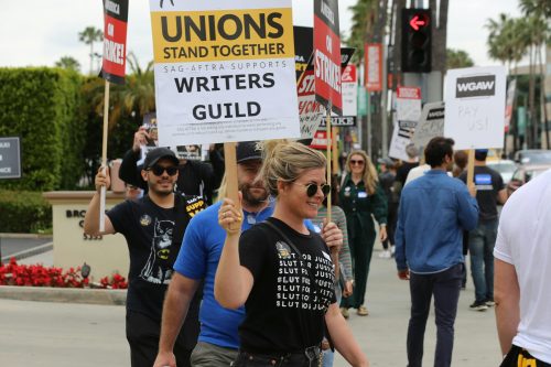 People holding signs and marching in a writers guild strike on a city sidewalk with palm trees in the background, spotlighting how the tech industry continues to impact the Business of Tech.