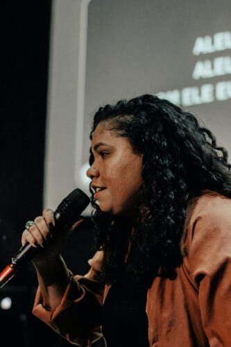 A woman with curly hair holding a microphone, singing or speaking on stage about the business of tech, with blurred text in the background.
