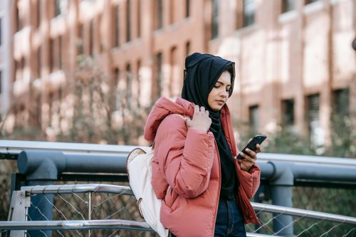 Woman in a pink jacket and black hijab stands on a bridge, immersed in her phone—a snapshot of today’s ever-evolving tech culture.
