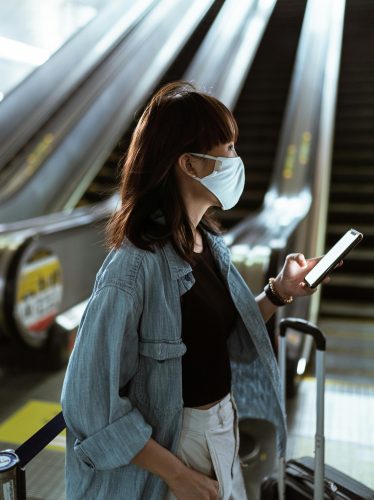 A woman wearing a face mask, holding a phone and suitcase, stands near escalators in an indoor setting—reflecting the impact of technology on modern travel and governance.