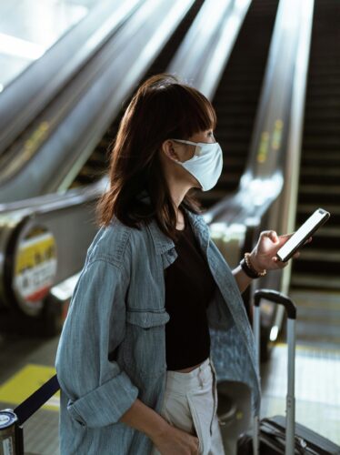 A woman wearing a face mask, holding a phone and suitcase, stands near escalators in an indoor setting—reflecting the impact of technology on modern travel and governance.