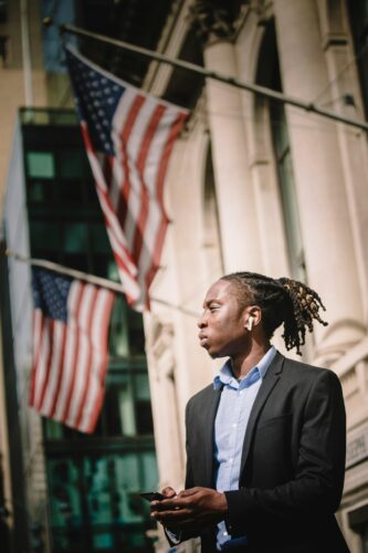 A man in a suit stands outside with two American flags and a tall building in the background, symbolizing leadership in technology governance.