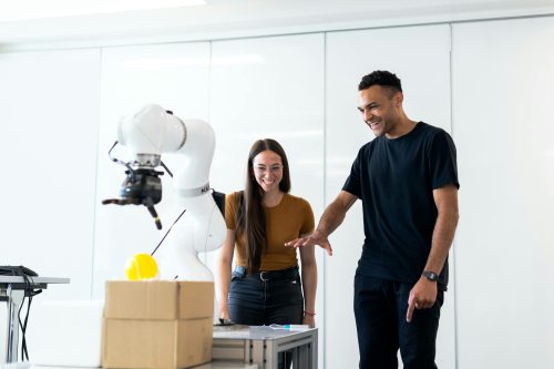 Two people smiling as they interact with a robotic arm on a table filled with boxes and objects, highlighting collaboration and innovation in the technology industry.