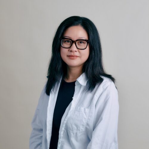 Woman with long dark hair and glasses, wearing a white shirt, sits against a plain light background.