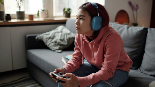 A person with headphones is immersed in video games, lounging on a cozy couch surrounded by lush plants.