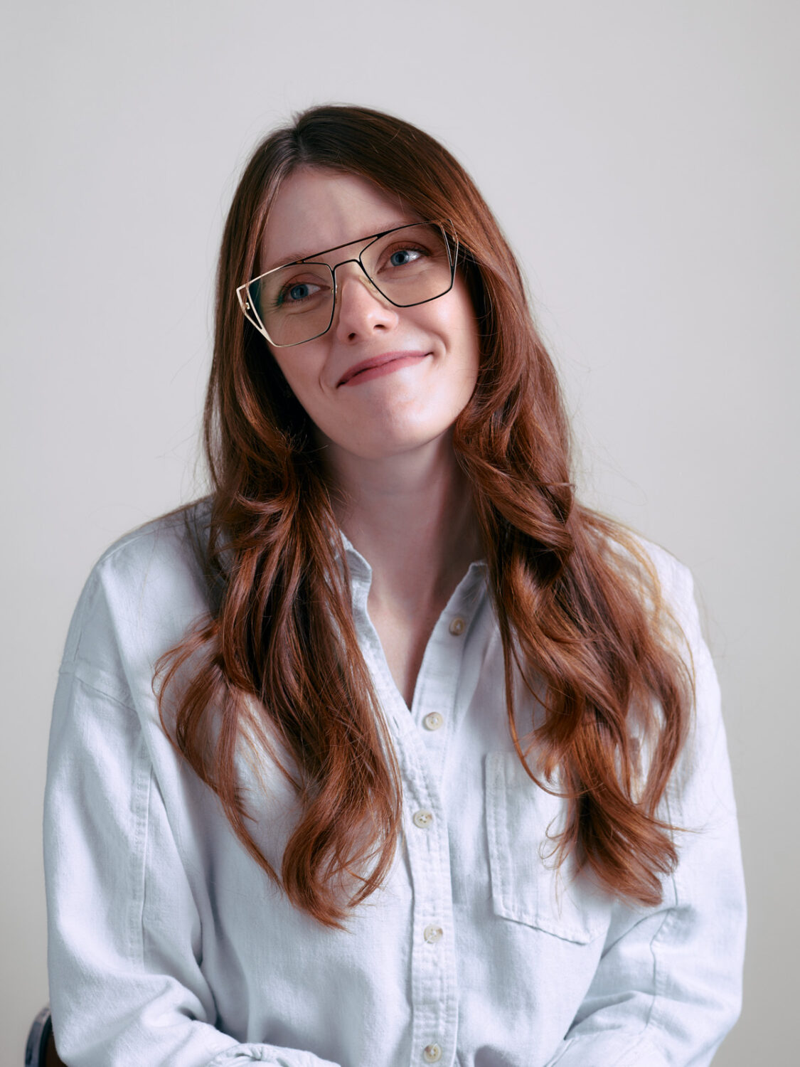 Woman with long brown hair and glasses in a white shirt, smiling softly against a light background, embodying the harmonious blend of AI innovation and everyday technology.