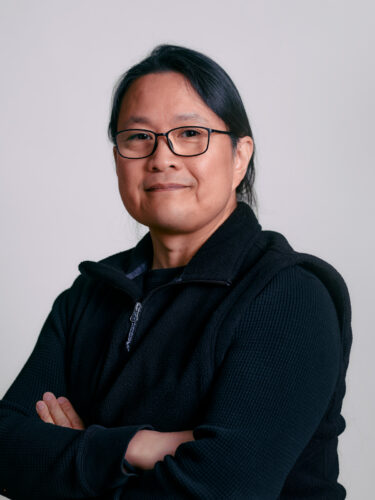 A person with glasses, long hair, and a black jacket smiles confidently with arms crossed against a plain background, embodying the spirit of responsible tech.