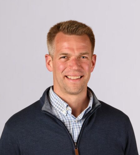 A smiling man in a blue sweater over a checked shirt stands against a plain white background, embodying the principles of responsible tech with his forward-thinking approach.