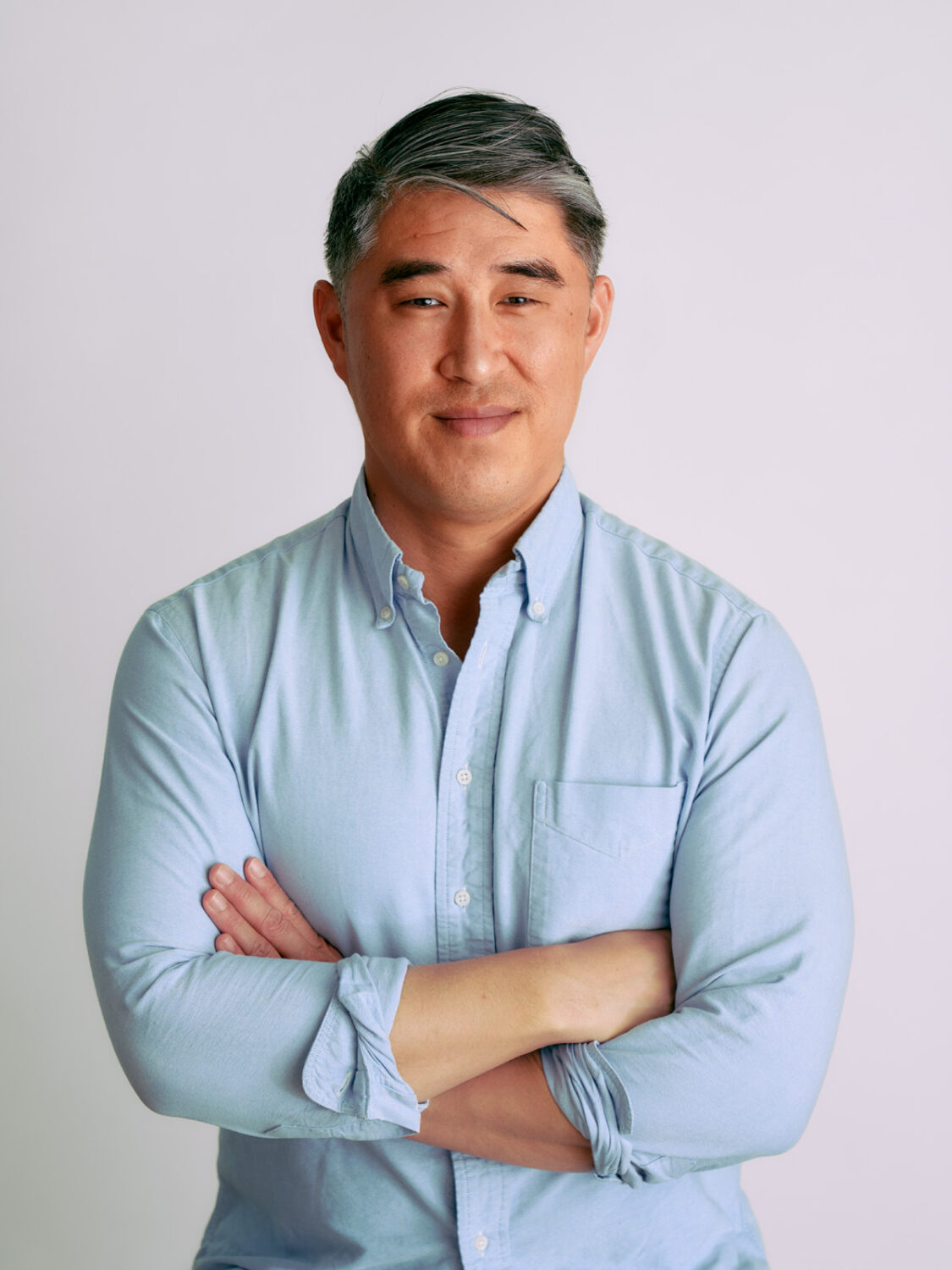 Smiling man in a light blue shirt poses with arms crossed against a plain white background