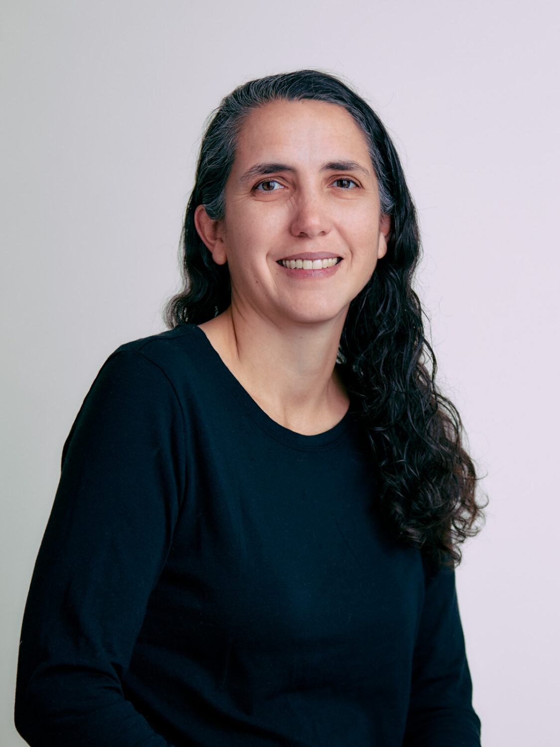 Gretchen Phillips, with long, wavy dark hair and a black shirt, smiles against a plain light background.