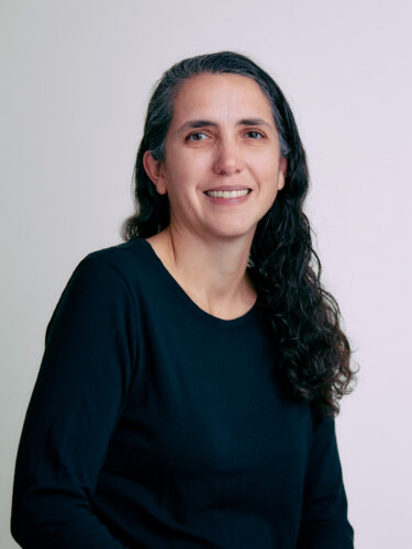 Gretchen Phillips, with long, wavy dark hair and a black shirt, smiles against a plain light background.