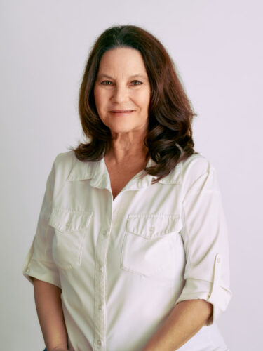 A woman with long dark hair wearing a white shirt, smiling against a plain background
