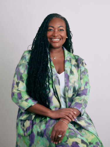 A smiling woman in a green and purple patterned suit sits with hands clasped in front, embodying the spirit of responsible tech that champions humanity.