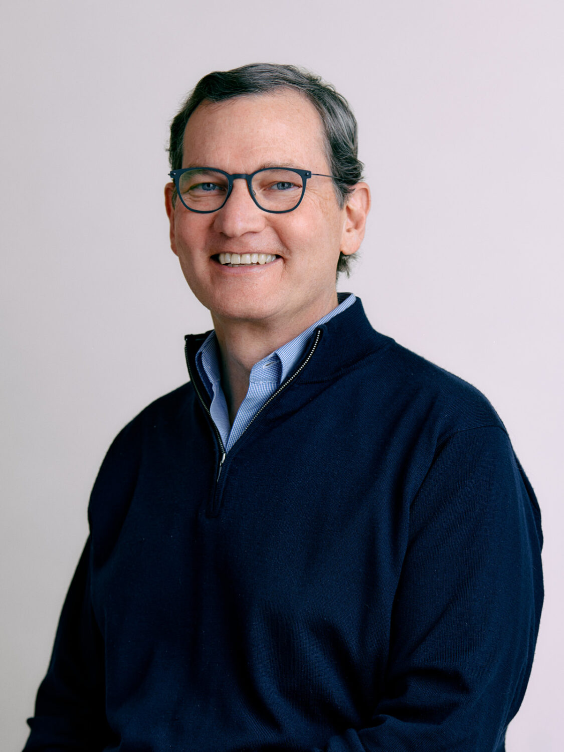 A person wearing glasses and a navy sweater is smiling warmly against a plain light background, embodying the seamless blend of technology in our everyday lives.