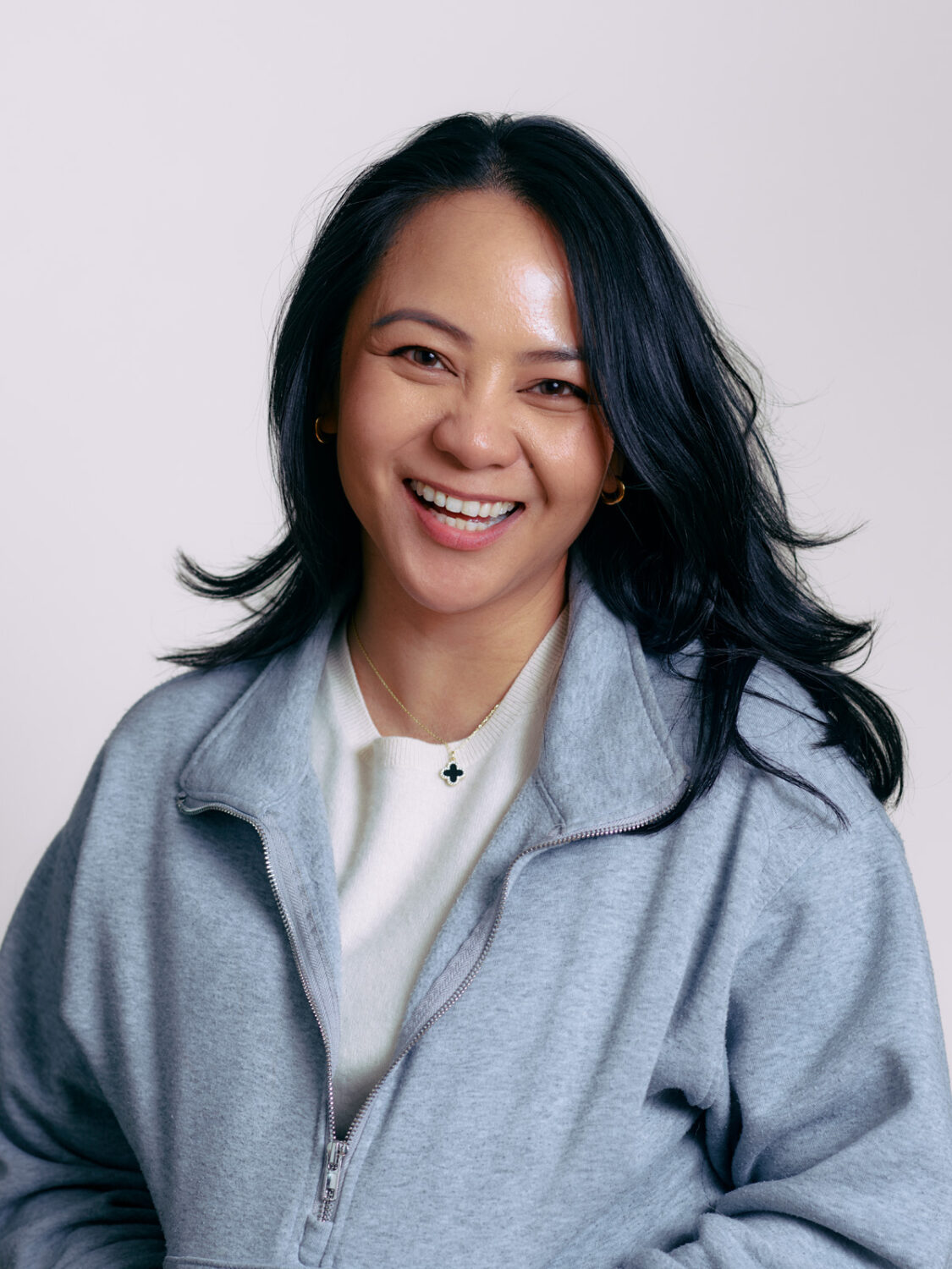 A smiling person with long dark hair, wearing a gray zip-up jacket and a necklace, stands against a light background