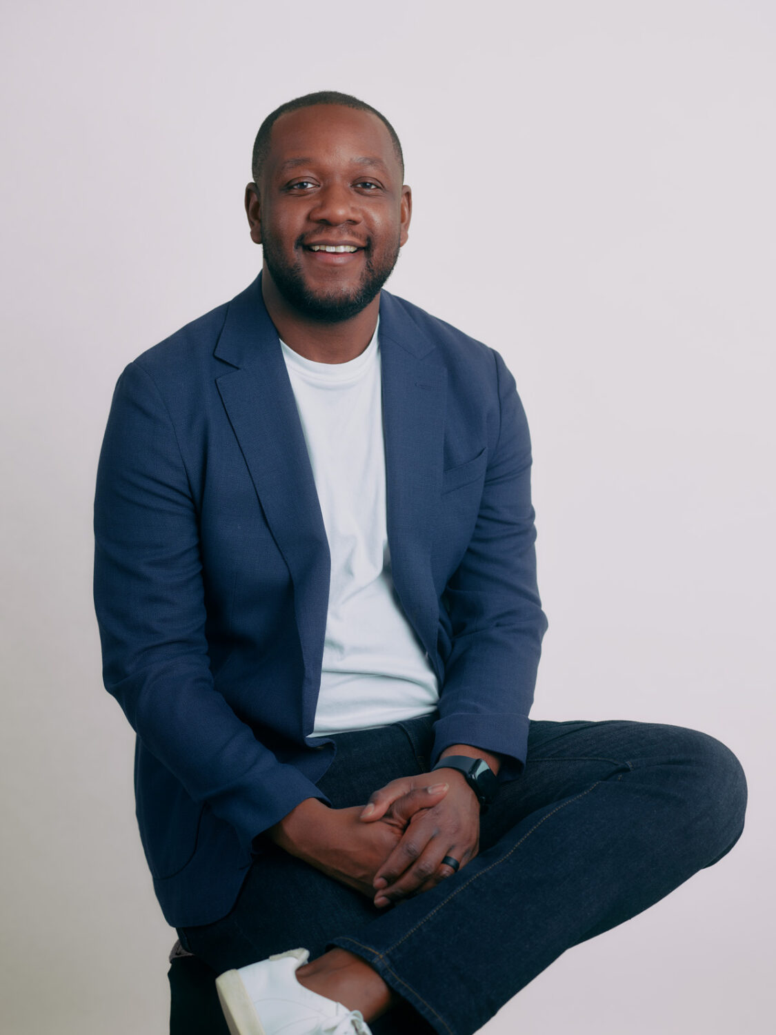 A person sits and smiles in a blue blazer, white shirt, and jeans against a plain backdrop