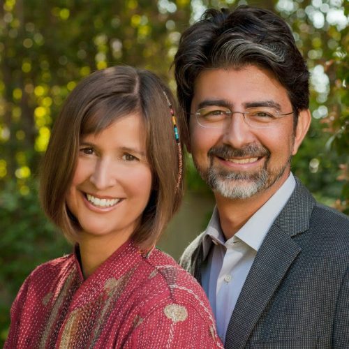 A smiling woman in a red patterned top stands next to a man in a suit, outdoors with greenery behind them.