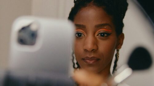 Person with braided hair uses a smartphone, wearing bright eye makeup, and holding a makeup brush.