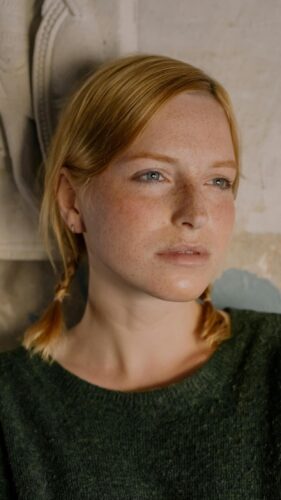 Woman with red hair in braids wearing a green sweater, standing against a textured wall.