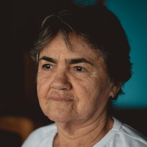 Older woman with short gray hair wearing a white t-shirt, looking pensively to the side.