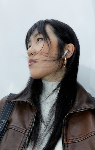 Woman with long black hair wearing a brown jacket and earbud, looking to the side against a white background.