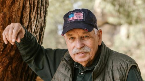 Elderly man in a cap and vest leaning against a tree outdoors, with farm equipment in the background.