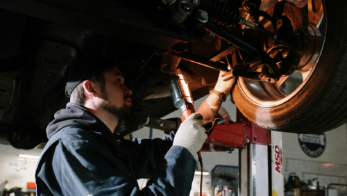 Mechanic working under a raised car, inspecting with a light and wearing gloves and a cap.
