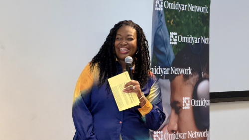 Michele L. Jawando, president of Omidyar Network, smiles while holding a microphone and notes in front of an Omidyar Network banner.