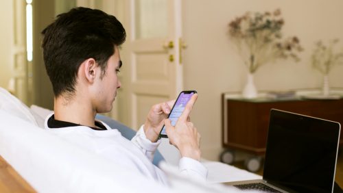Person sitting on a couch using a smartphone, with an open laptop nearby on a table in a cozy room, reading about the recent Apple antitrust ruling.