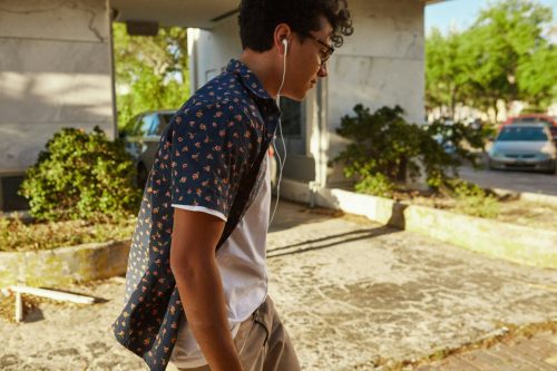Young man with curly hair and earphones, possibly a sixth class student, walking outside on a sunny day, wearing a patterned shirt.
