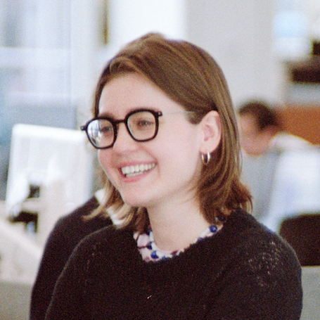 Smiling person with glasses holds a plate with pizza slices in an office setting.