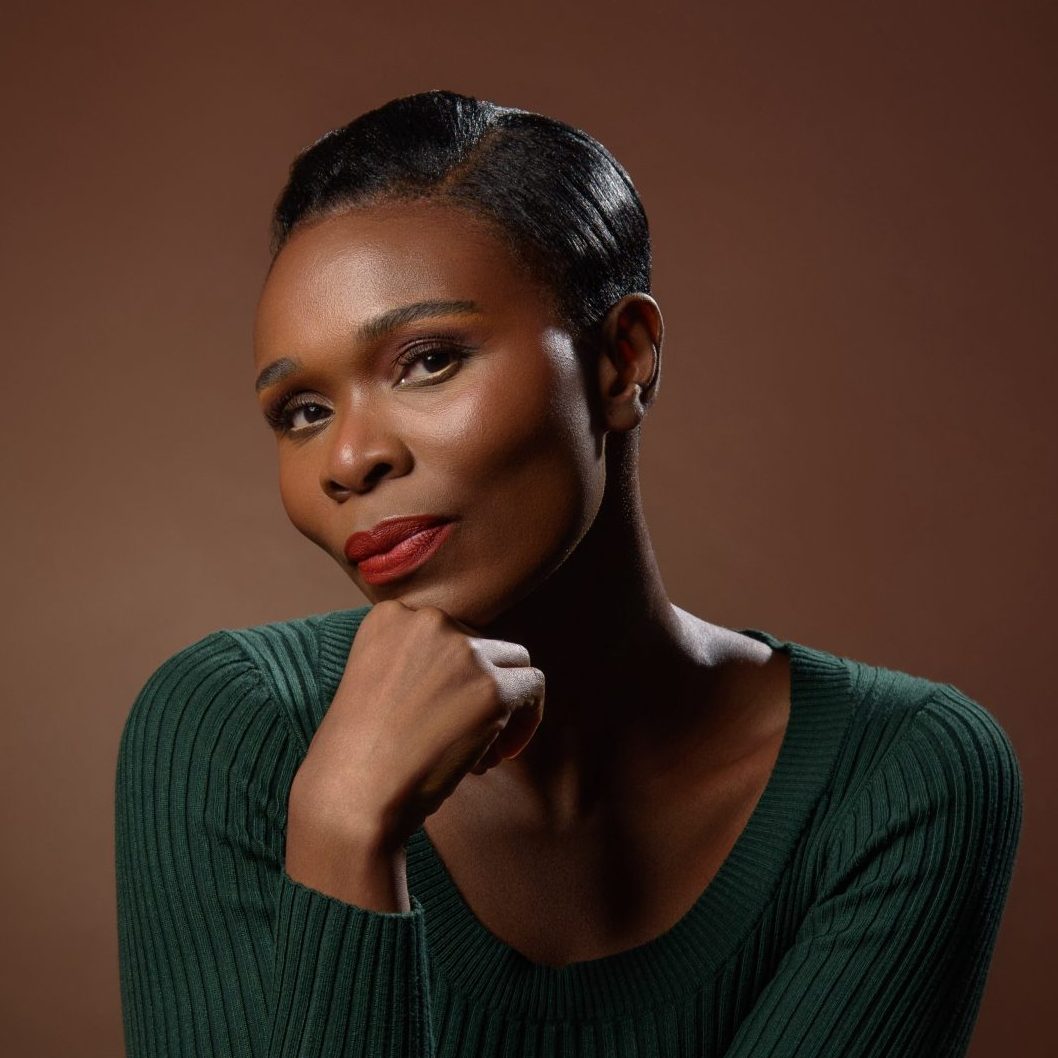 Woman in a green sweater poses with chin resting on her hand against a brown background, looking thoughtful.