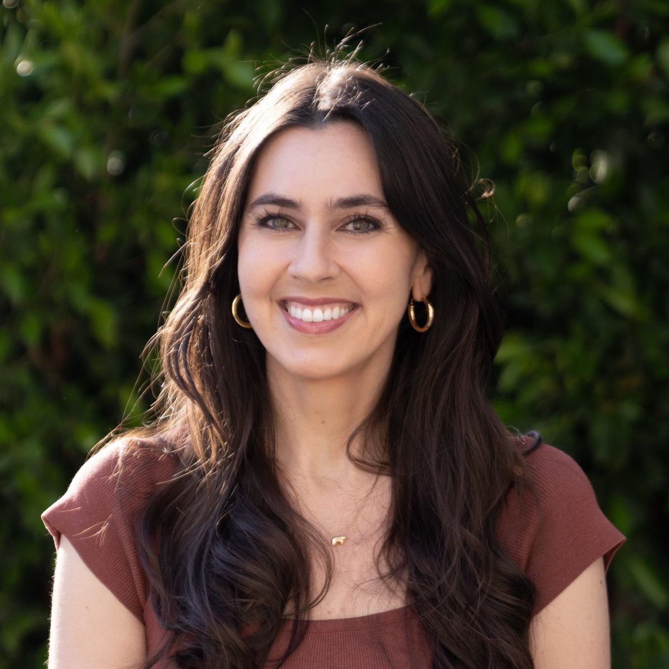 Woman with long brown hair wearing a brown top smiles outdoors in front of green leafy bushes.