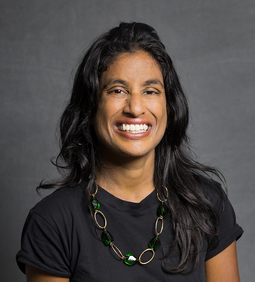 Woman with long dark hair, smiling, wearing a black shirt and a necklace, in front of a gray background.