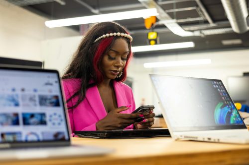 Woman in a pink blazer uses her phone at a desk with two open laptops, possibly researching topics like the Legislative Academy on AI and how California policymakers are advancing AI in an office setting.