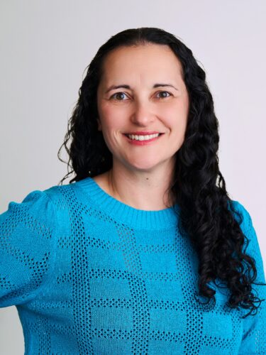 Smiling woman with long curly dark hair in a bright blue sweater, standing against a light background.