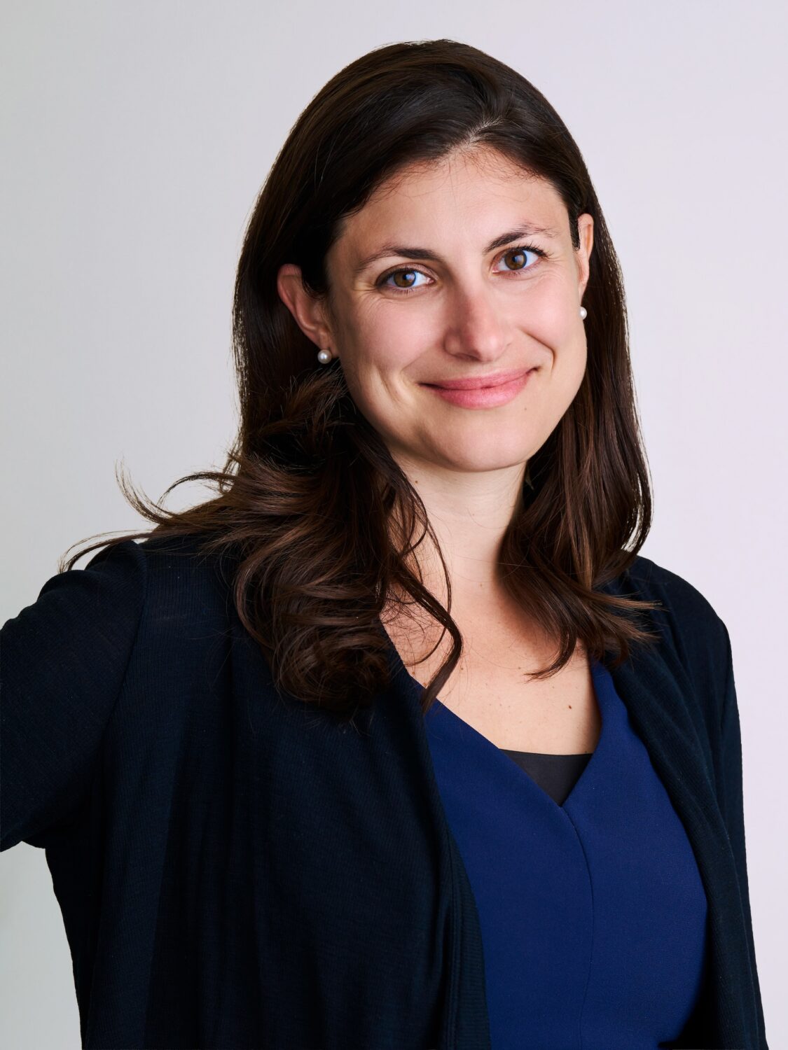 Smiling woman with long brown hair in a navy blue top and black cardigan against a light background.