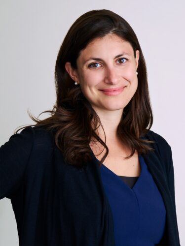 Smiling woman with long brown hair in a navy blue top and black cardigan against a light background.