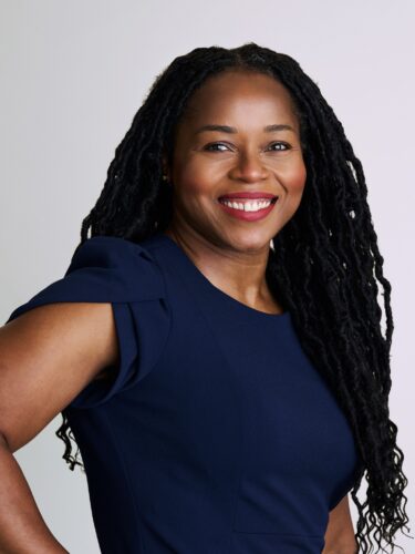 Smiling woman with long braided hair wearing a navy blue dress, standing against a plain light background.