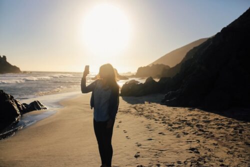 Person standing on a sandy California beach at sunset, holding up a phone with rocky cliffs in the background.