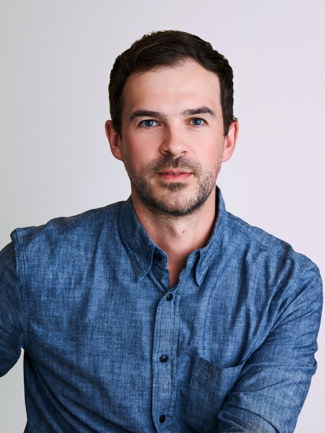 Man with short dark hair and beard wearing a blue button-up shirt, posed in front of a plain background.